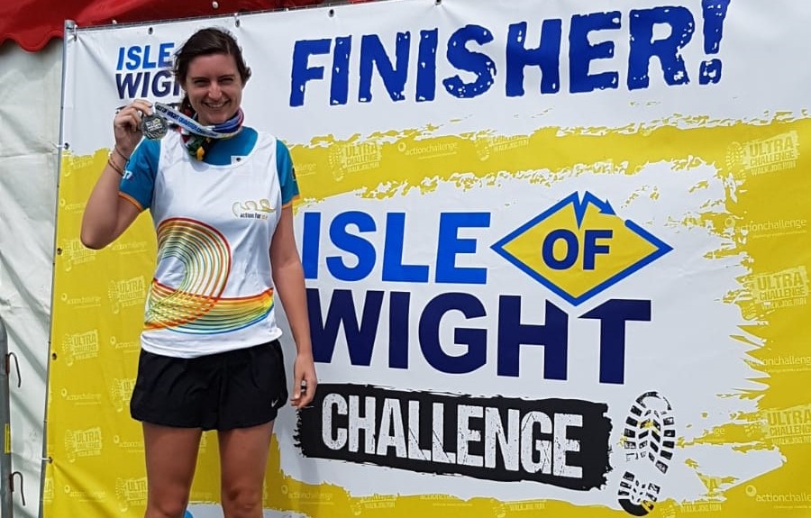A person smiles, holding up an Isle of Wight Challenge finishers medal, wearing an Action for ME running vest. They are stood in front of a yellow sign reading 