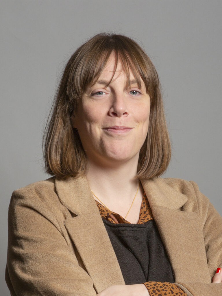 A woman with shoulder-length brown hair wearing a beige coat over a black top poses against a grey background, arms crossed.
