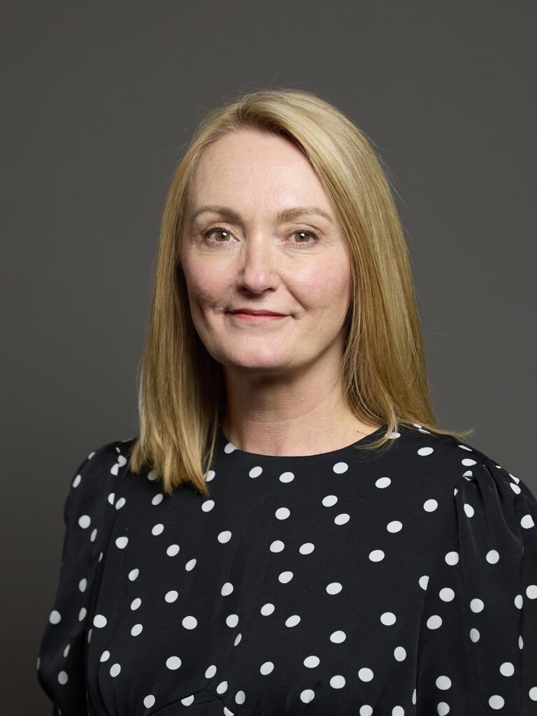 A portrait of a woman with long blonde hair wearing a black polka dot blouse against a grey background.