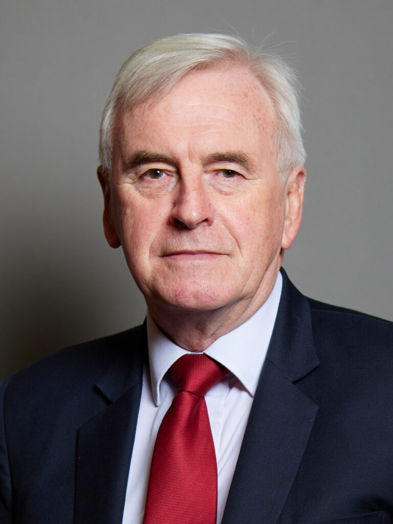 A man in a dark suit and red tie poses against a neutral background.