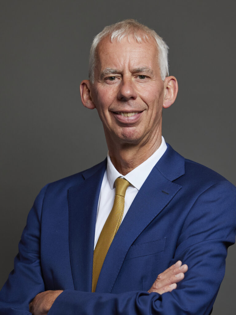 A formal portrait of John Milne MP, smiling in a blue suit with a yellow tie.