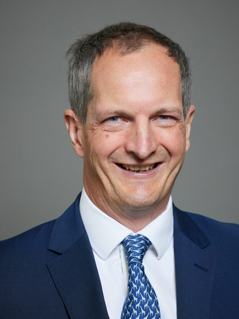 A person wearing a navy suit and a patterned tie stands against a plain grey background.