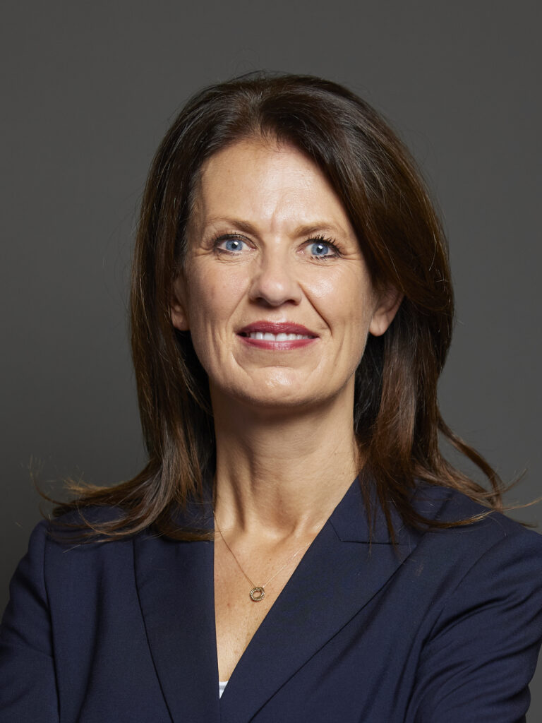 A woman with long brown hair wears a navy blazer and a delicate necklace, standing against a dark grey background.