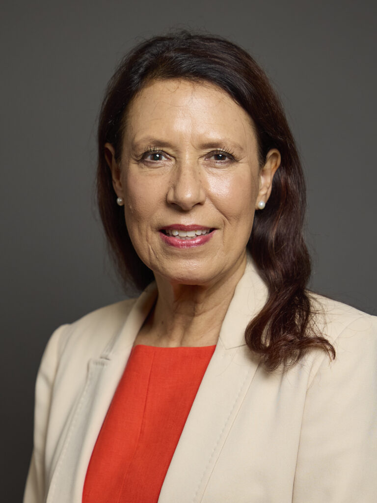 A woman in a cream blazer and red top is posed against a grey background.