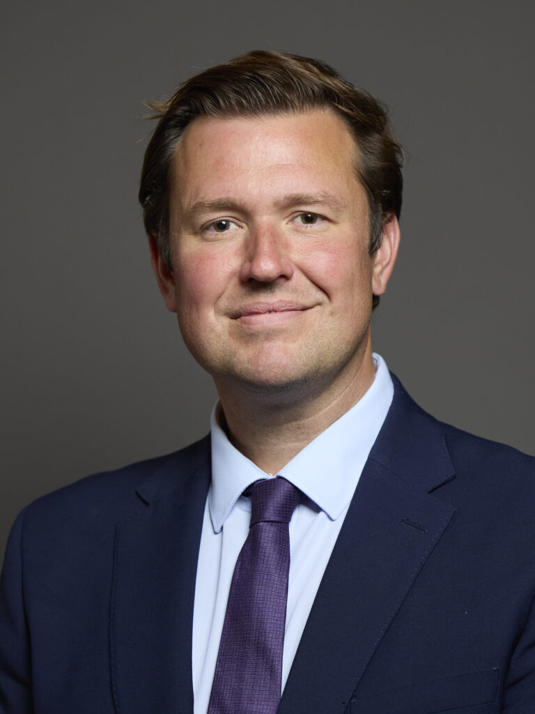 Portrait of a man in a dark suit with a light blue shirt and purple tie, set against a neutral grey background.