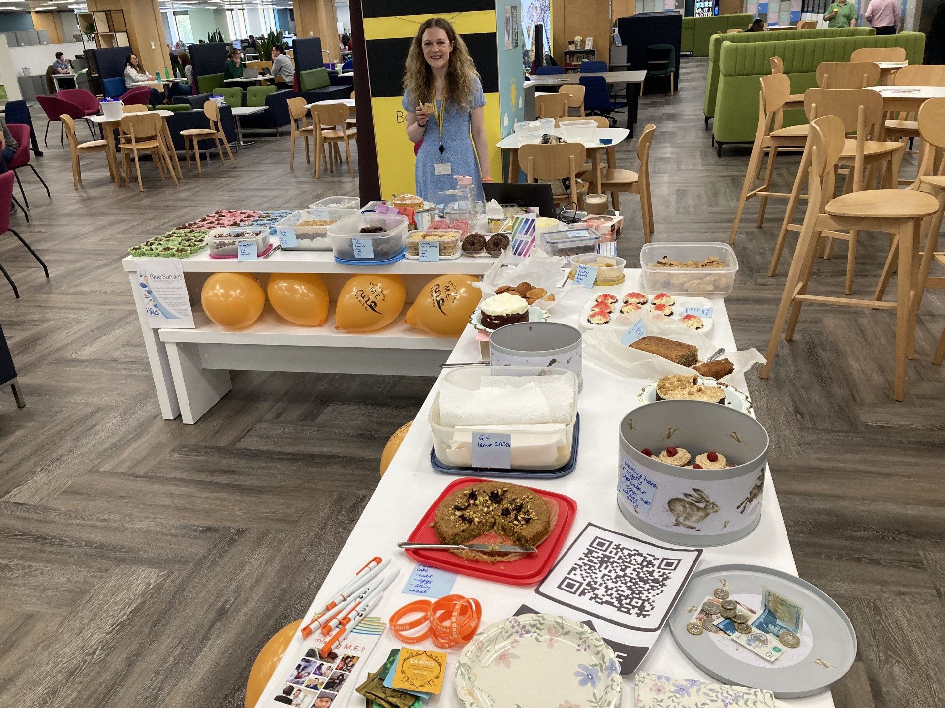 A woman with long hair stands, smiling at the camera, in front of an l-shaped table full of cakes and baked goods. Action for ME and Blue Sunday-related merchandise is also laid out across the table.
