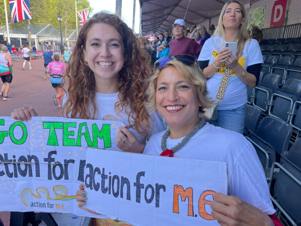 Two Action for ME staff wearing Action for ME t-shirts, cheering on London Marathon runners, holding signs reading 