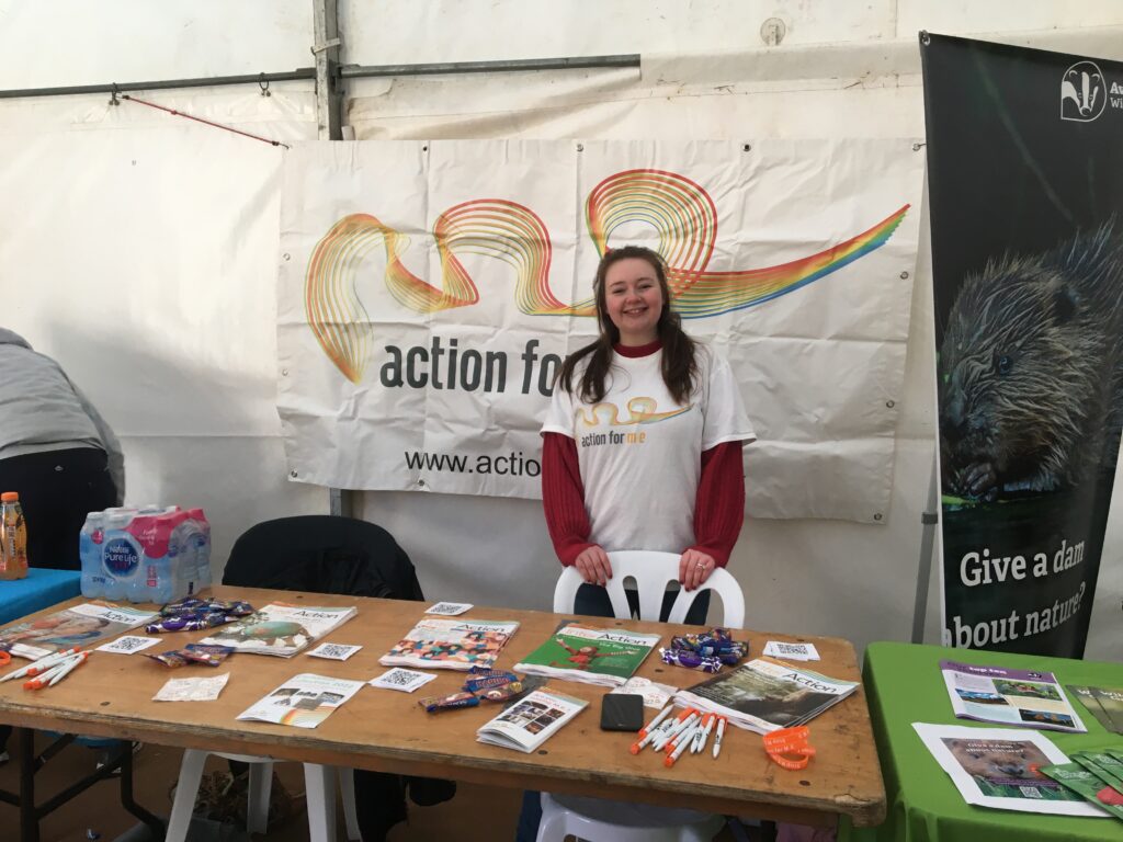 An person standing in front of a table covered in Action for ME fundraising materials. They are stood in front of a big banner with Action for ME's logo on it.