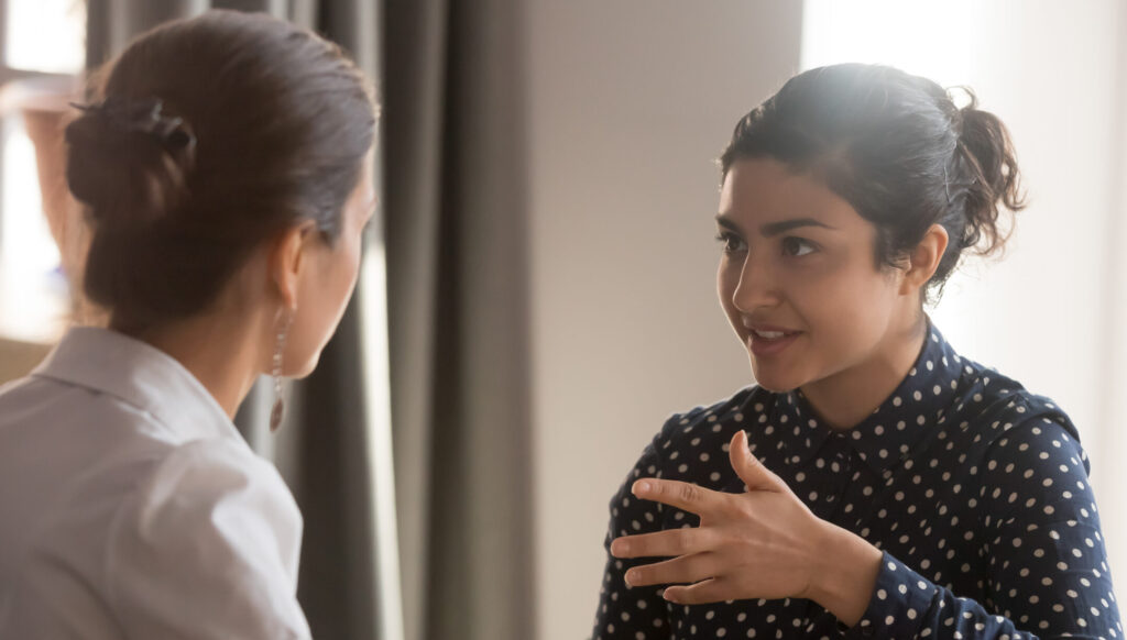 Two women engage in discussion, seated in a softly lit room with curtains and warm decor. One gestures expressively.