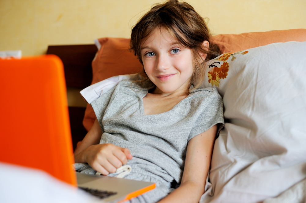A child relaxes in bed with a laptop, wearing a grey shirt and surrounded by pillows, looking at the viewer.