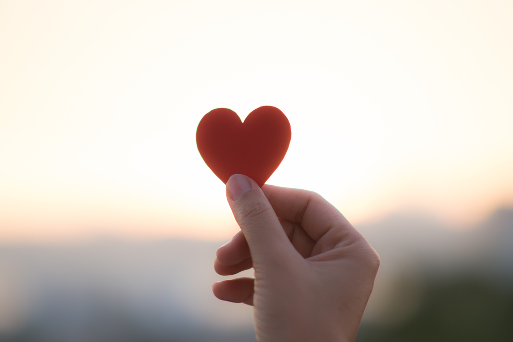 A hand holds up a small, red, heart-shaped piece of card, against a blurred sunset.