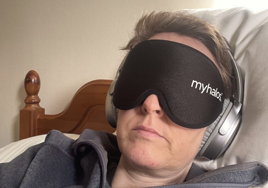 Lisa, who has ME, lying down wearing a pair of noise-cancelling headphones and a MyHalos eye mask.