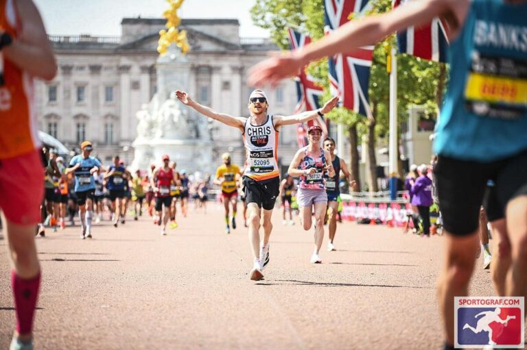 A group of runners participating in a race near Buckingham Palace. The central runner, wearing a bib labeled 'CHRIS' with number '52095,' has arms raised in celebration. Union Jack flags line the route, and other runners are visible in the background. The logo 'SPORTOGRAF.COM' appears in the bottom right corner.