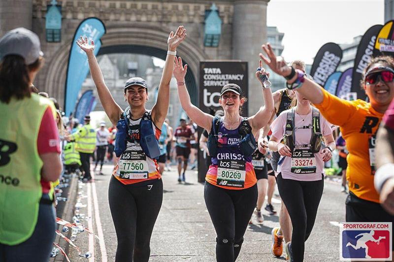 A group of marathon runners pass under an archway during a race event, with some raising their arms in celebration. Visible bib numbers include 77560, 80269, and 33274. Banners and signs in the background feature branding such as 'TATA CONSULTANCY SERVICES' with the TCS logo. Spectators and event staff line the sides of the path