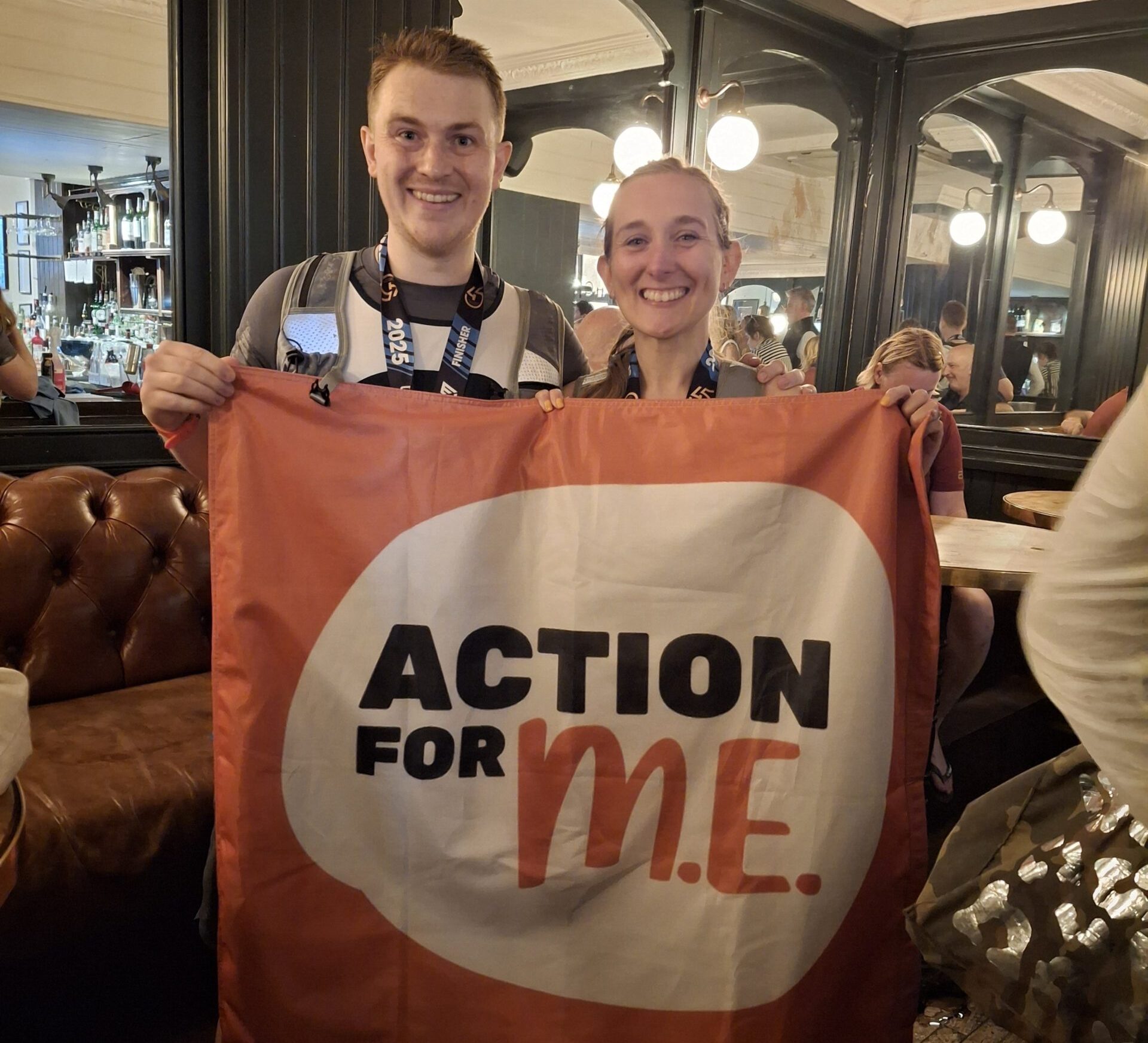 Two Action for M.E. fundraisers holding a banner that reads 'ACTION FOR M.E.'.The photo is taken in a restaurant or café setting, with tables, chairs, and lights in the background
