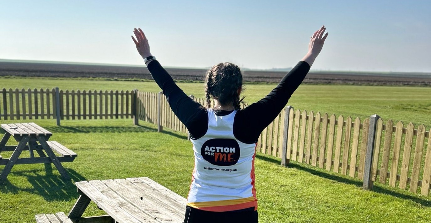 A person with braided hair is seen from behind standing on a grassy field with arms raised in celebration. They are wearing a white running vest with black sleeves that reads 