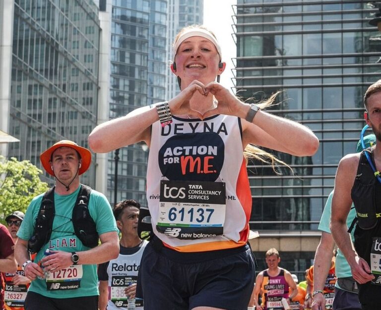 A group of marathon runners is seen participating in a city race surrounded by tall modern buildings. The central runner, a smiling woman wearing a headband and a jersey labeled "DEYNA" and "Action for M.E.," is making a heart shape with her hands. She has the race number 61137. Other runners, both male and female, wear various athletic gear and bib numbers. The atmosphere is energetic and sunny, with spectators and greenery visible in the background.