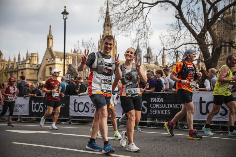 A group of marathon runners is participating in a race, with some making peace signs towards the camera. The runners are wearing numbered bibs and athletic gear. In the background, a crowd of spectators stands behind barriers with banners displaying 'TCS' and 'Tata Consultancy Services.' The setting appears to be an urban area with historic buildings and a church spire visible in the distance.