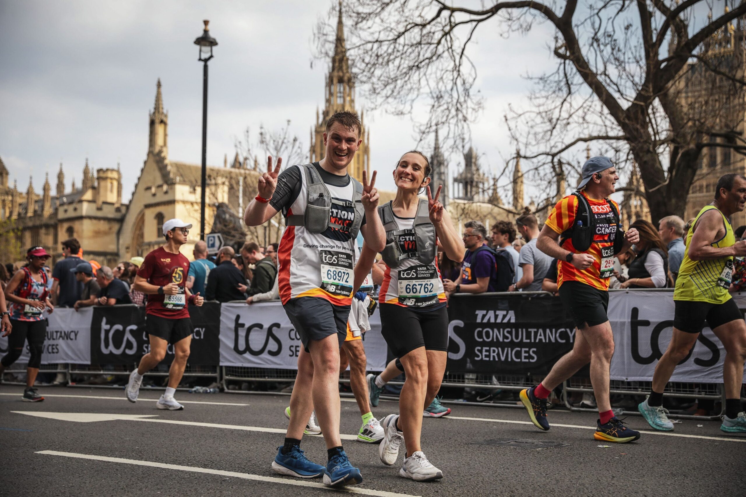 A group of people, including Action for M.E. fundraisers, participating in a marathon or running event. They are running on a road lined with barriers, behind which a crowd of spectators is gathered. Some runners are making peace signs with their hands. The background features historic buildings and urban architecture, indicating the event is taking place in a city with notable landmarks