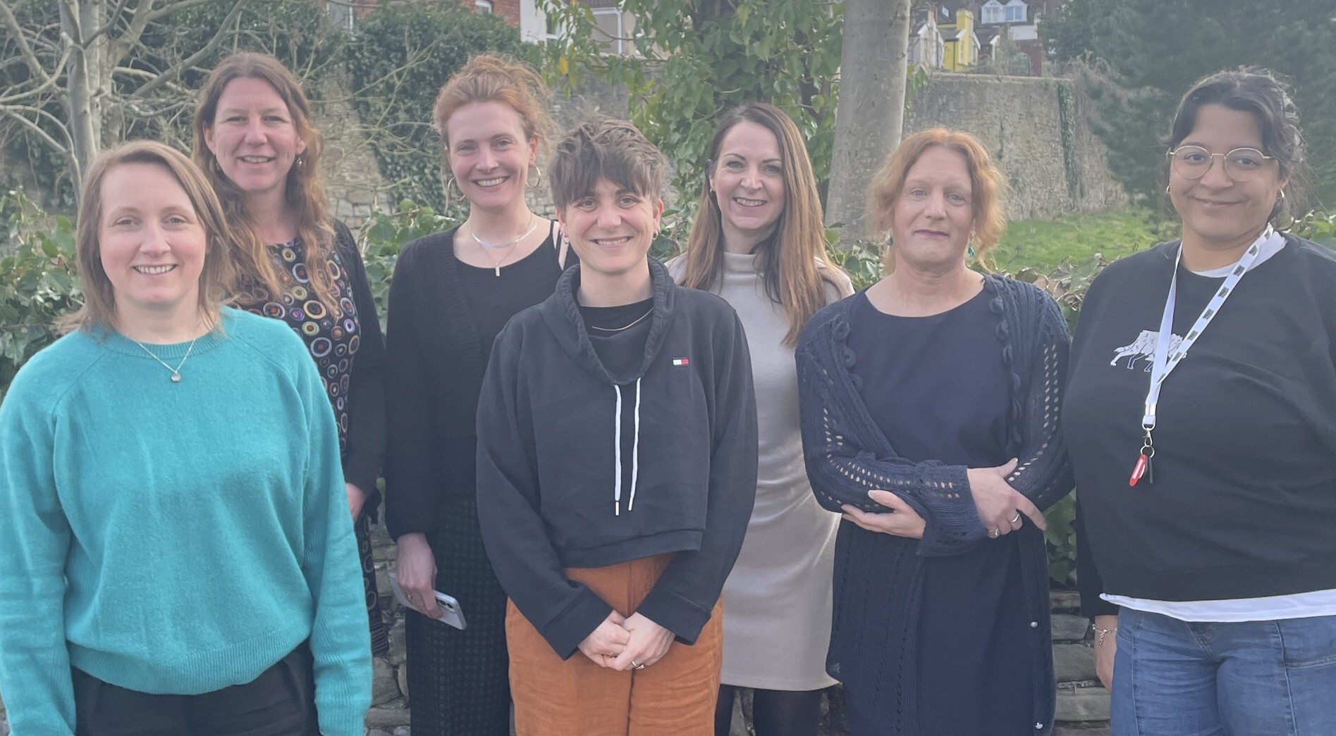 A group of seven people - the Action for ME fundraising team - standing outdoors in front of a tree and greenery. The sky is partly cloudy, and houses are visible in the background. The individuals are dressed in casual clothing, including sweaters, dresses, and jackets.