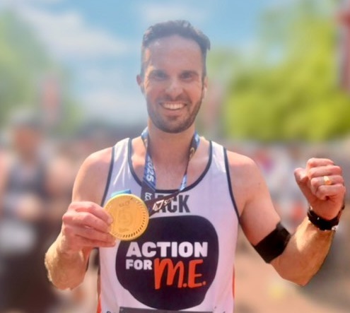 A smiling man stands proudly after completing a race, holding up a finisher’s medal in one hand and making a fist with the other. He is wearing a running vest with the logo 