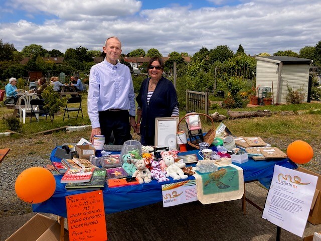 A man and a woman stand behind a table at an outdoor event. The table is covered with various items for sale, including books, stuffed animals, mugs, and other small goods. Two orange balloons are attached to the table. In the background, people are sitting at tables near a garden area with greenery and sheds.