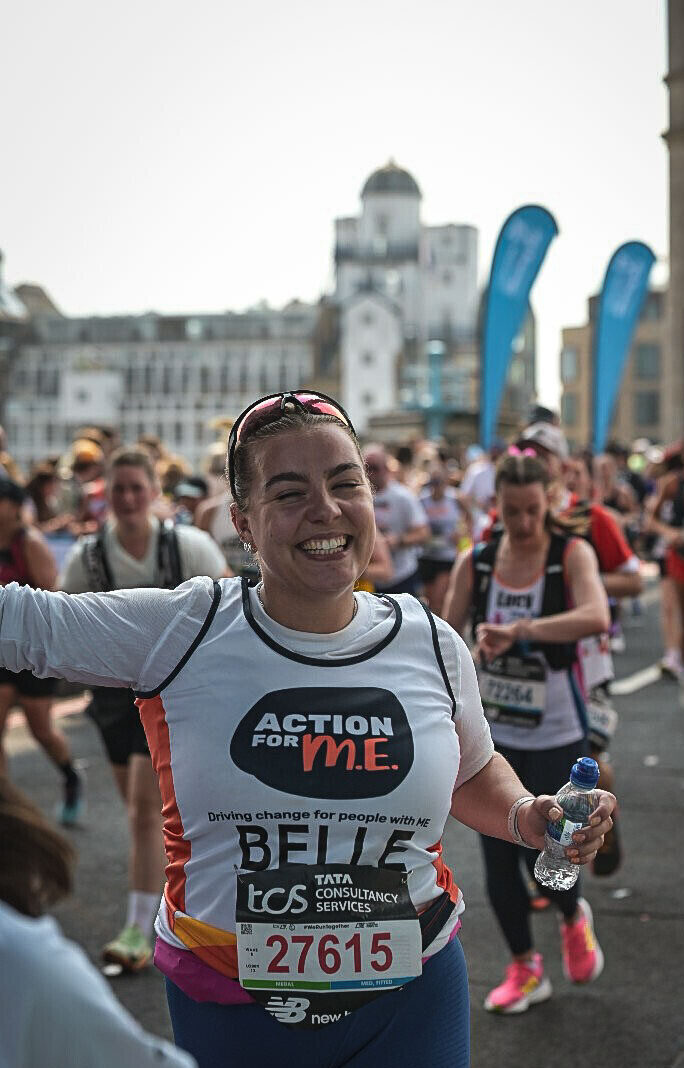 A group of runners participating in a marathon event. The central runner wears a white and black shirt with the text "ACTION FOR M.E." and "Driving change for people with M.E." Their bib number is 27615, featuring "TCS" and "BELLE." Other runners and buildings with blue flags are visible in the background, and some participants are holding water bottles.