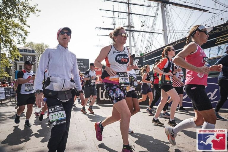 A group of runners participating in a marathon event, with some wearing bib numbers and athletic gear. In the background, a large sailing ship is docked at a pier, with trees and buildings visible to the left. The image is taken during daylight, and the runners are in motion.
