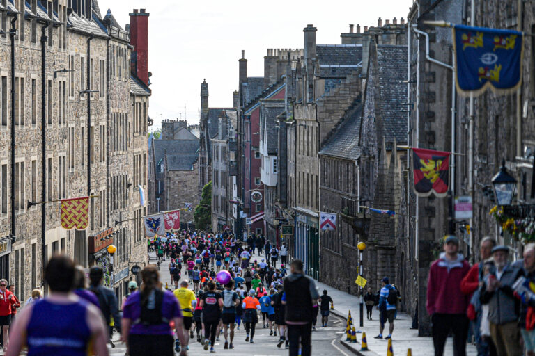 A bustling street scene shows a large group of people participating in a marathon or running event. The street is lined with historic stone buildings adorned with colorful banners and flags. Participants are dressed in various athletic attire, some wearing bib numbers, and are running or walking down the slightly inclined road. Spectators stand on the sidewalks, watching and cheering on the runners. The sky is overcast, casting a soft light over the scene.