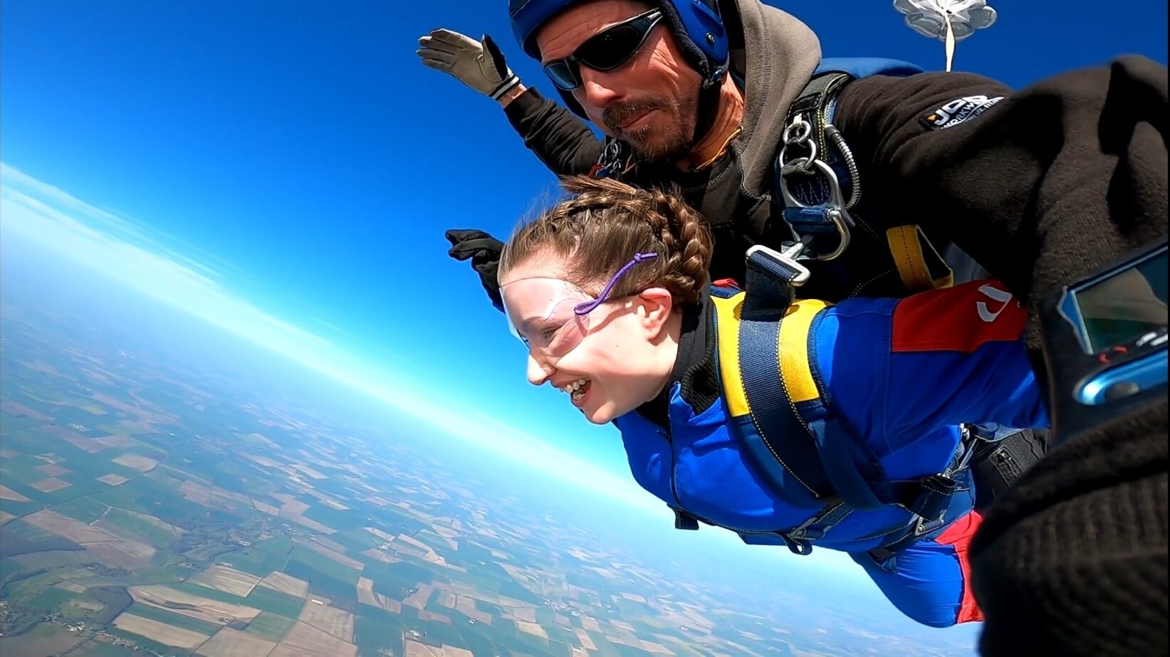 Person in a blue jumpsuit tandem skydiving with an instructor, freefalling above patchwork fields under a clear blue sky.