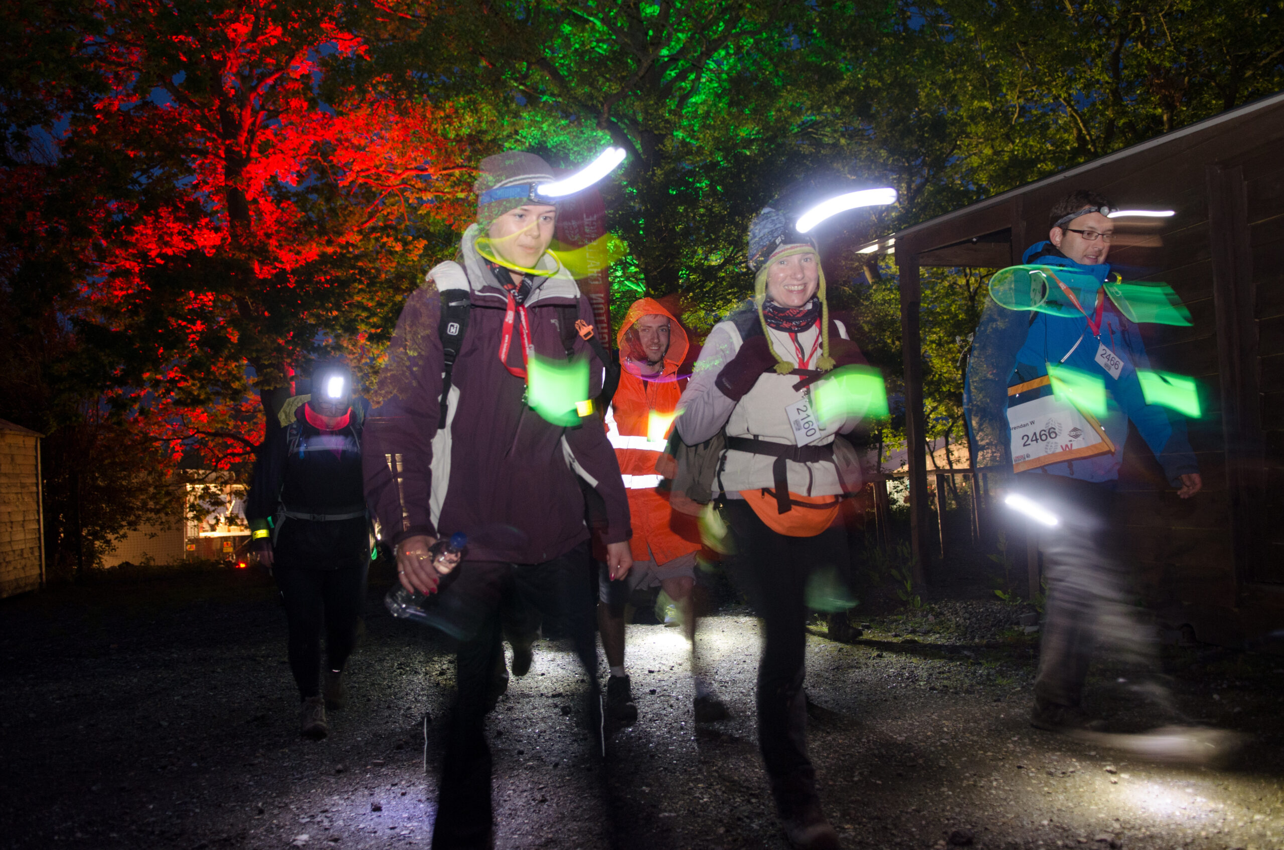 Group walking on a dark trail with headlamps and glow sticks, trees lit in red and green.