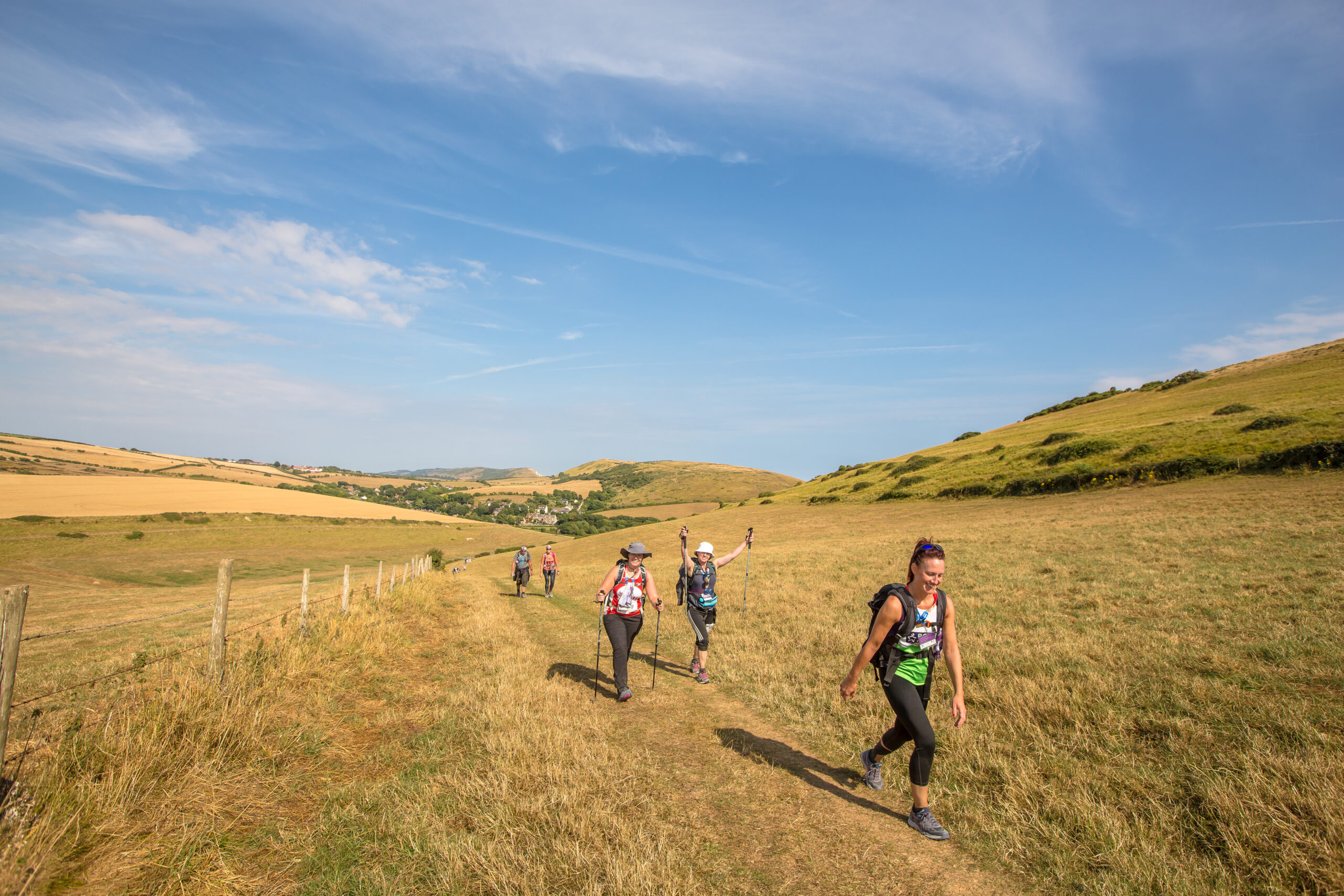 People walking along a grassy trail through open rolling hills under a clear blue sky.
