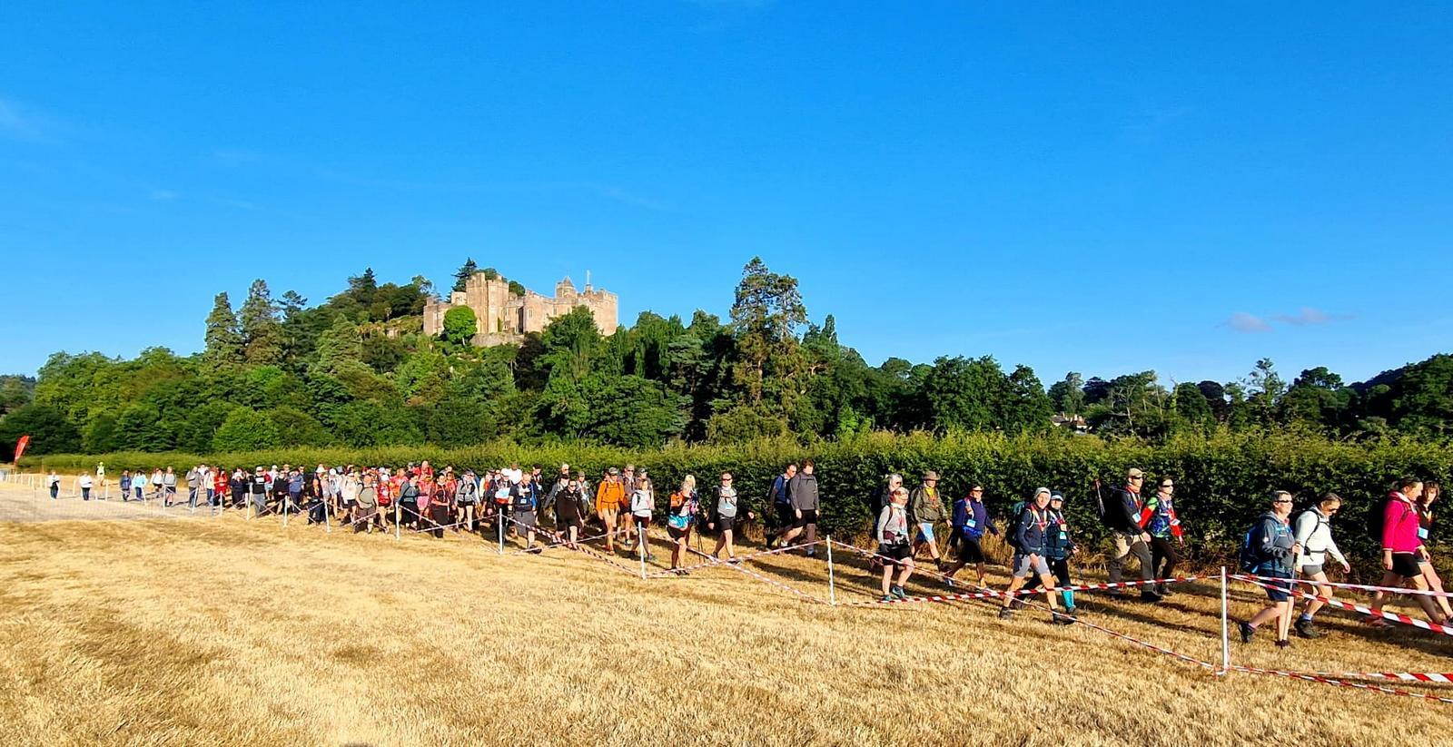 Large group of walkers on a grassy path with a castle and trees in the background under a clear blue sky.
