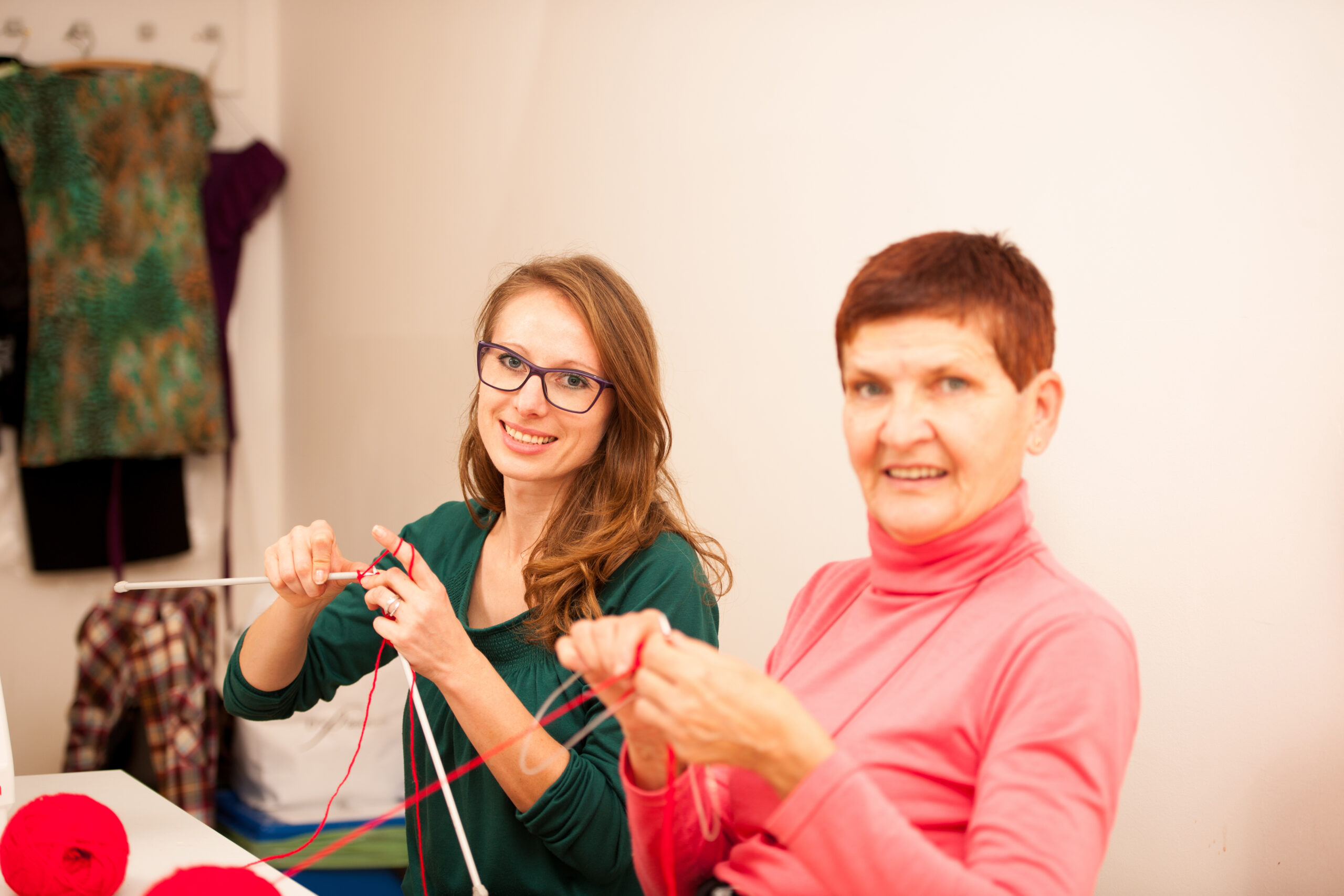 Two people sitting together indoors, knitting with red yarn.