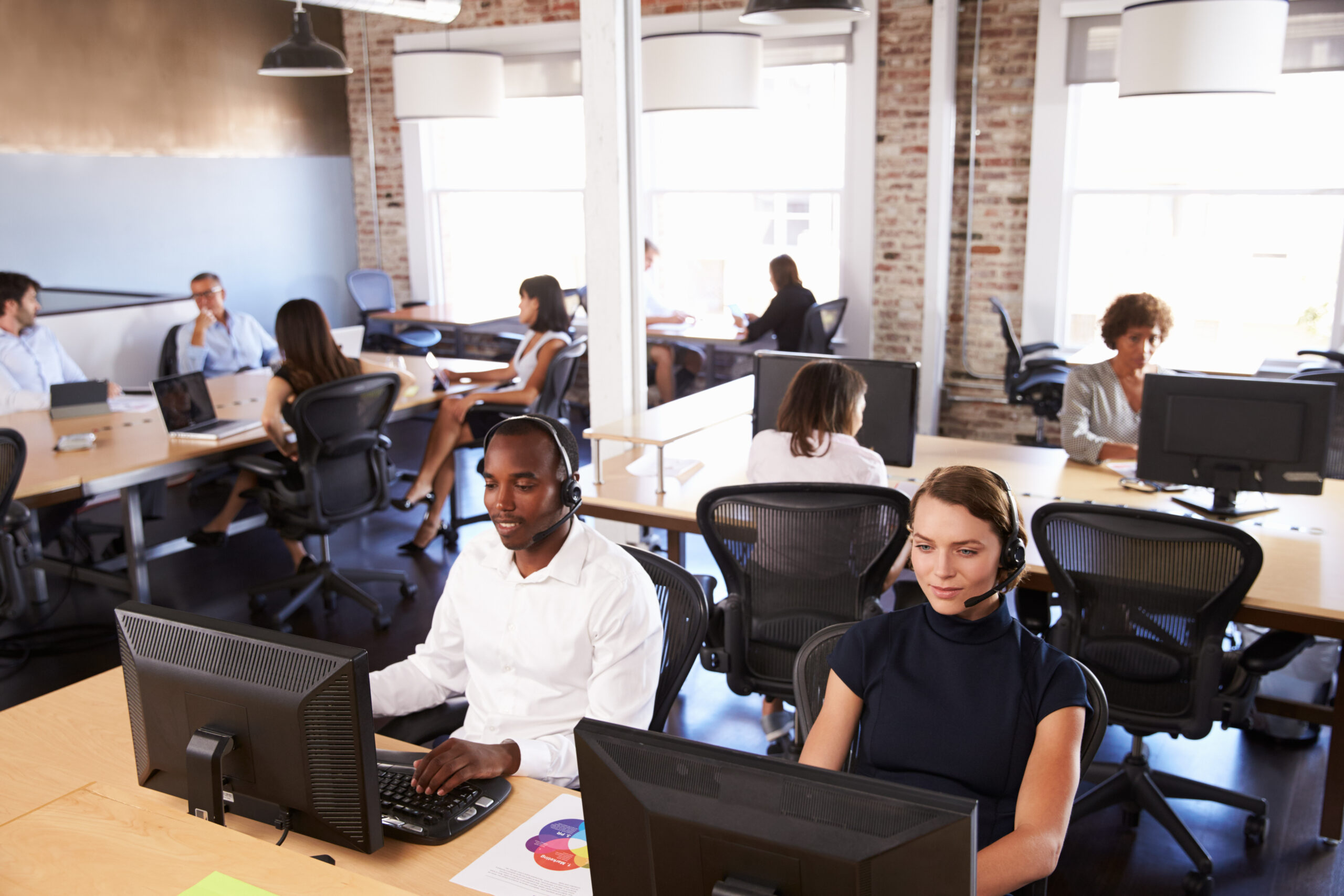 People working at desks in a bright, open-plan office with computers and headsets