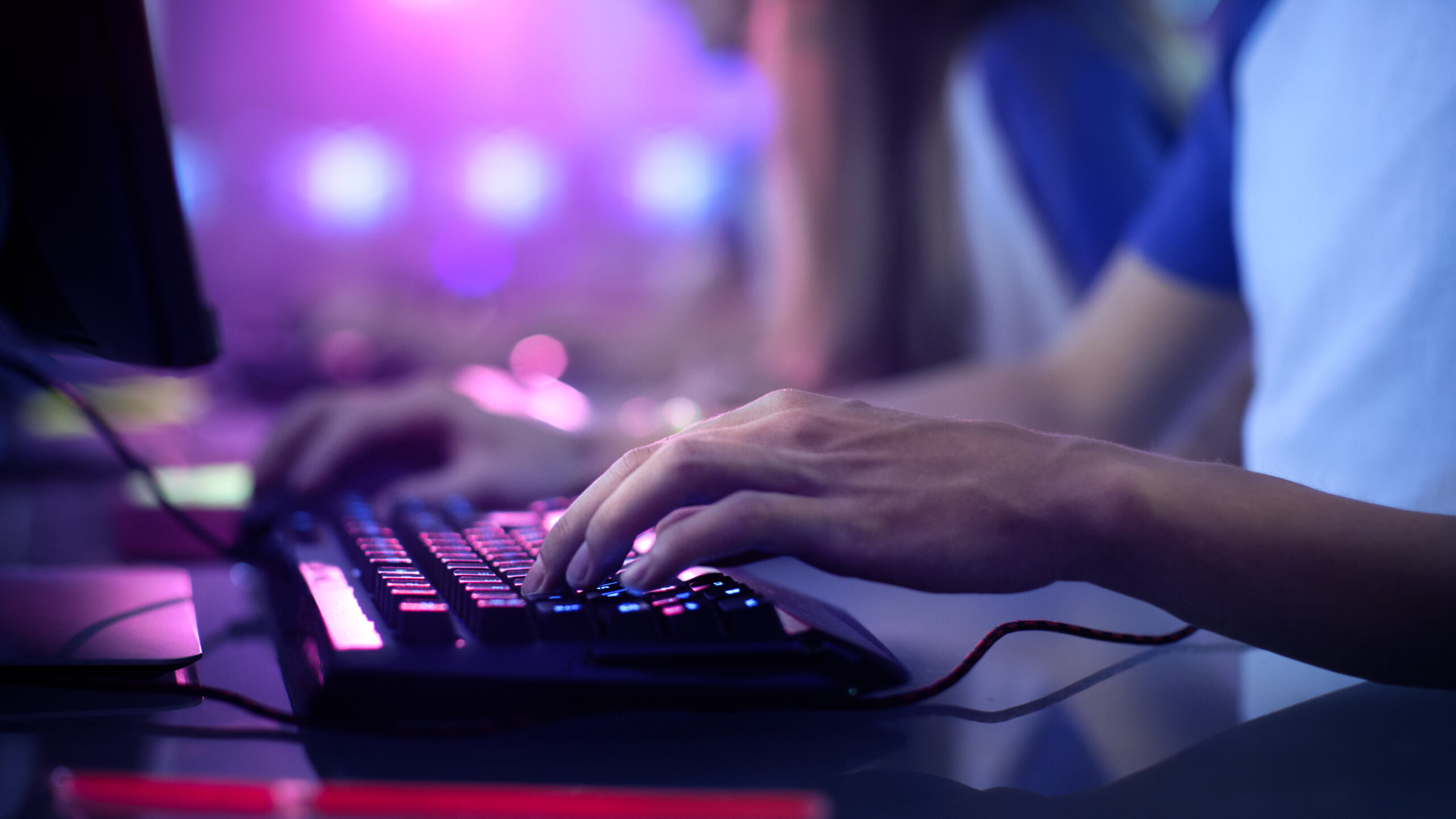 Hands typing on a backlit computer keyboard in a dim, colourful gaming environment.
