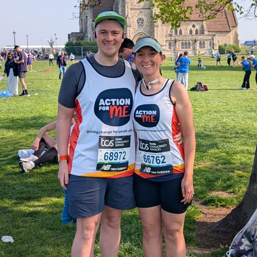Two people standing side by side under a tree in a park, wearing athletic gear and bibs with numbers 68972 and 67672. The bibs display the text 'ACTION FOR M.E.', indicating their participation in an event.